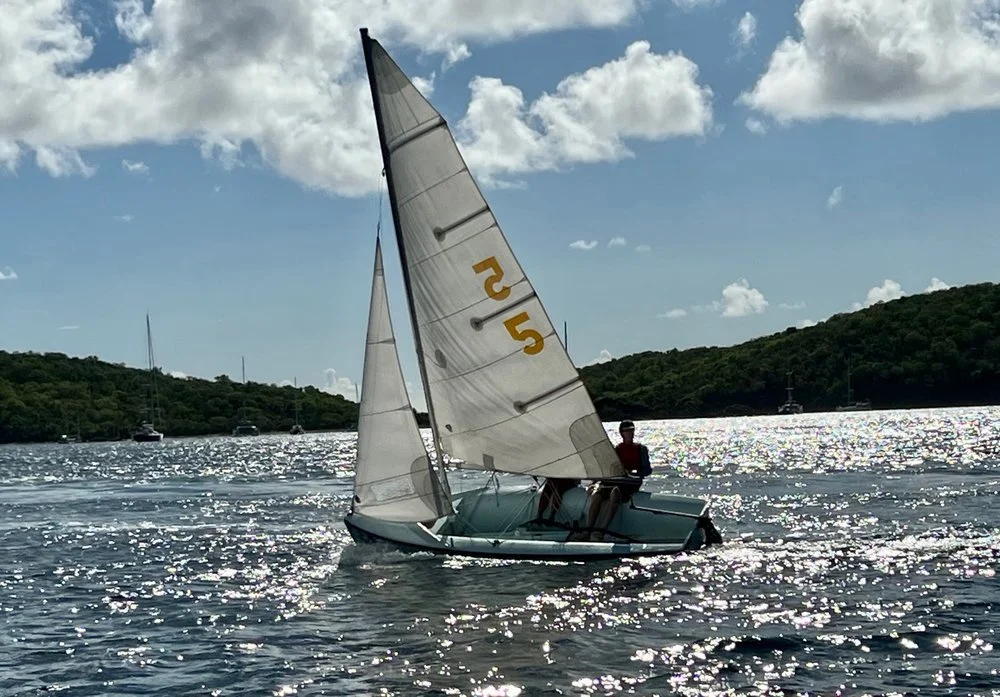 A person sailing a small boat with white sails on a sparkling body of water under a partly cloudy sky with green hills in the background.