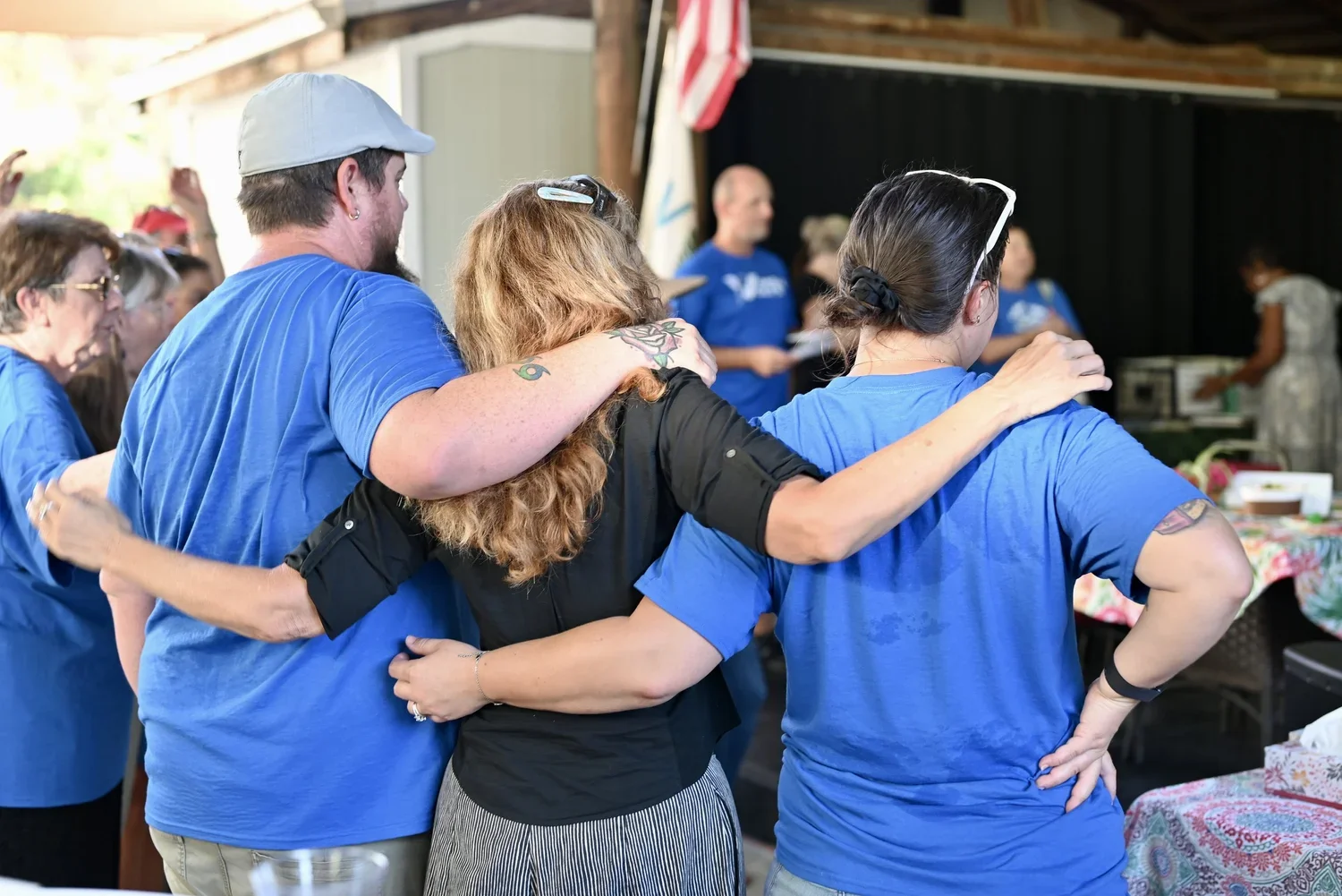 People participating in a group hug or prayer circle, with arms around each other's shoulders, wearing casual clothing and gathered indoors or at an event.