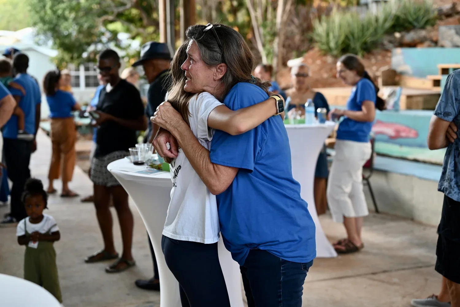 Two women hugging at an outdoor gathering, with other people in the background.