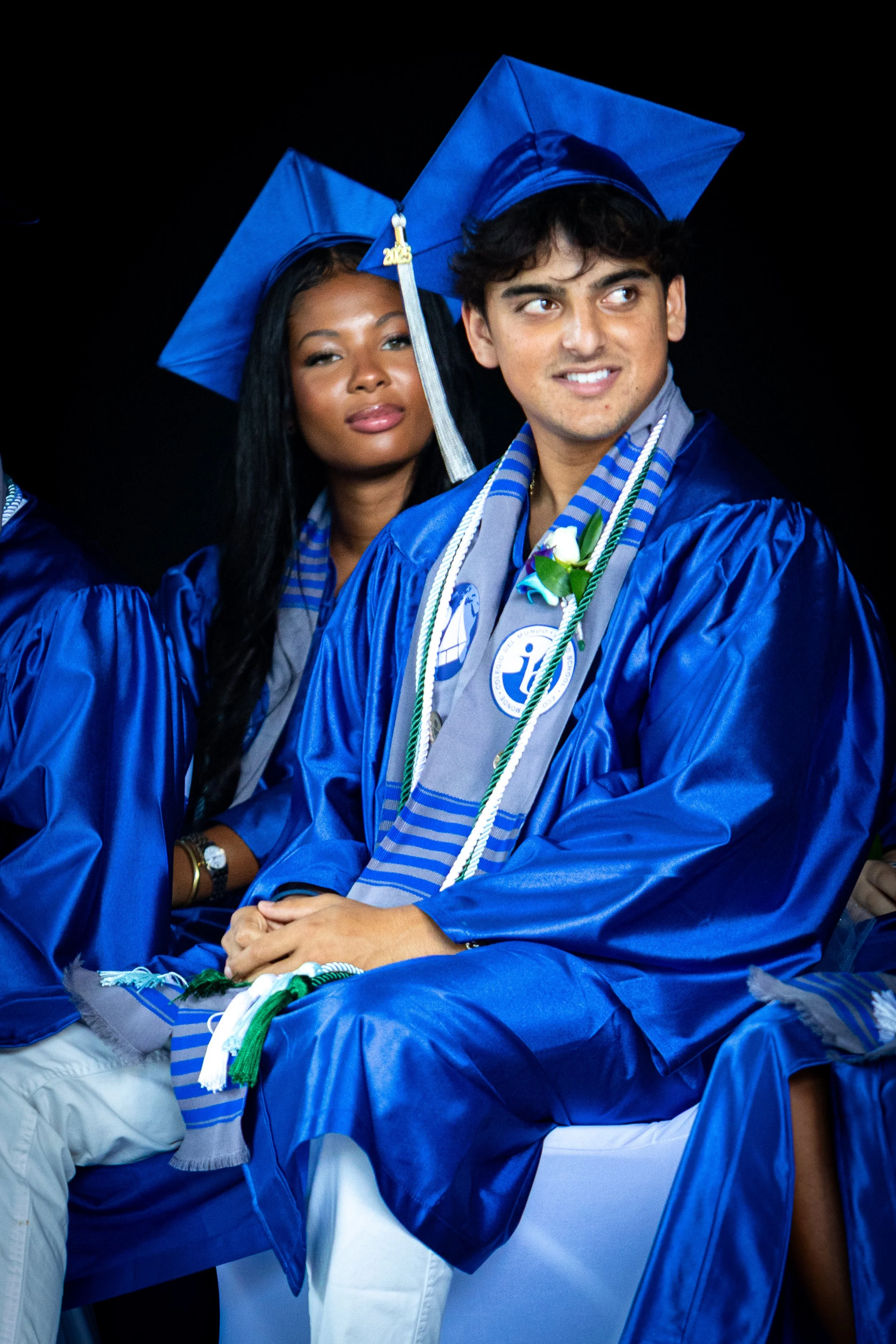 Two young graduates in blue caps and gowns, sitting at a graduation ceremony, with one smiling and the other looking serious.