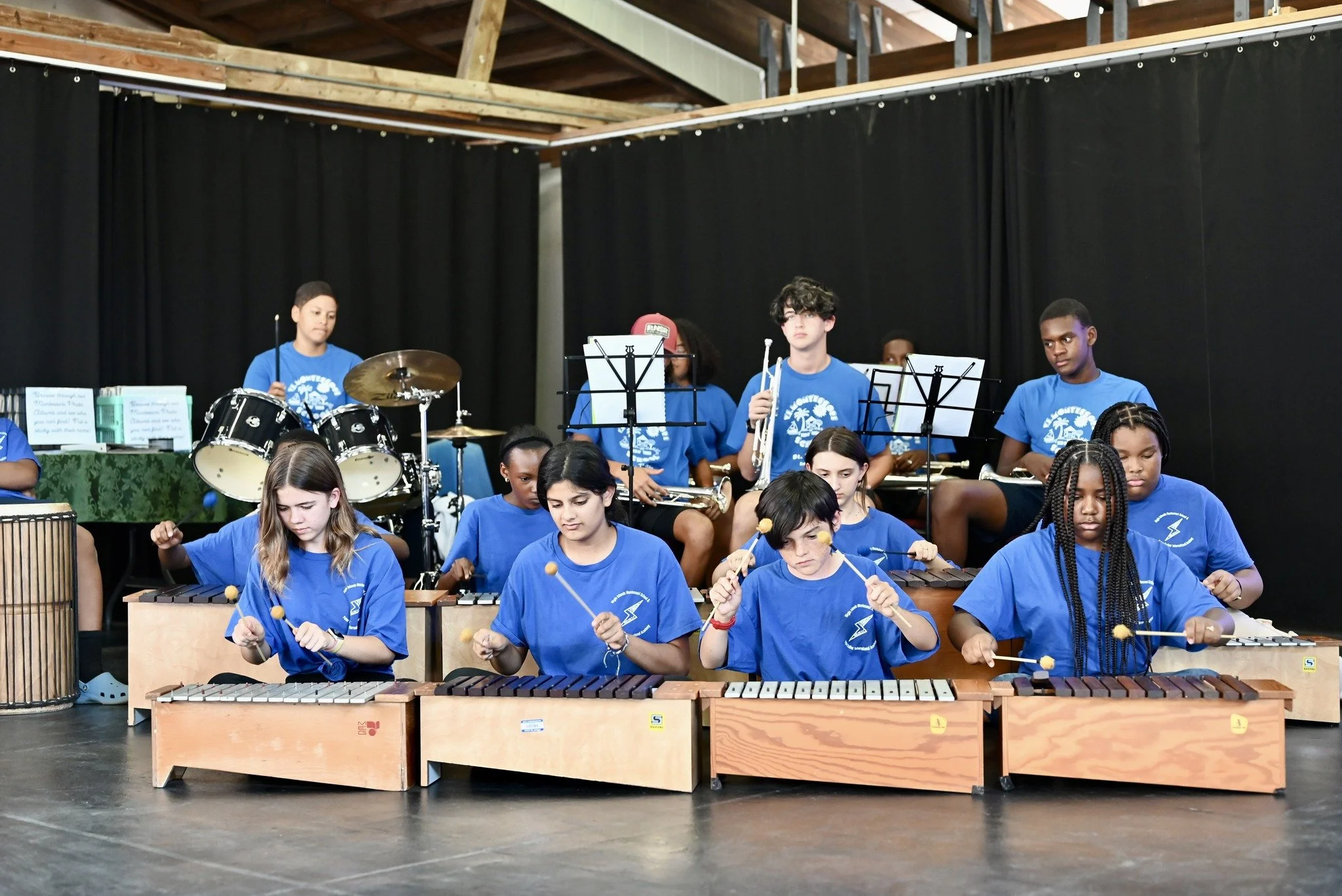 Group of young people playing musical instruments on stage, including xylophones, drums, and brass instruments, wearing matching blue t-shirts.