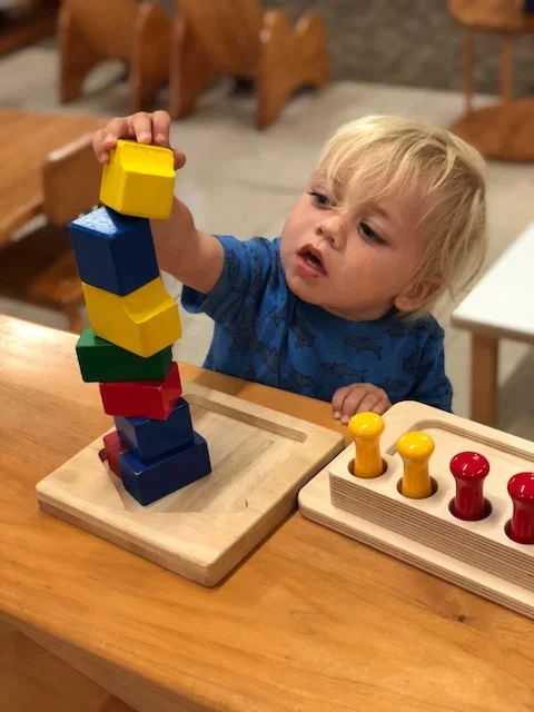 A young child stacking colorful wooden blocks on a wooden table in a playroom.