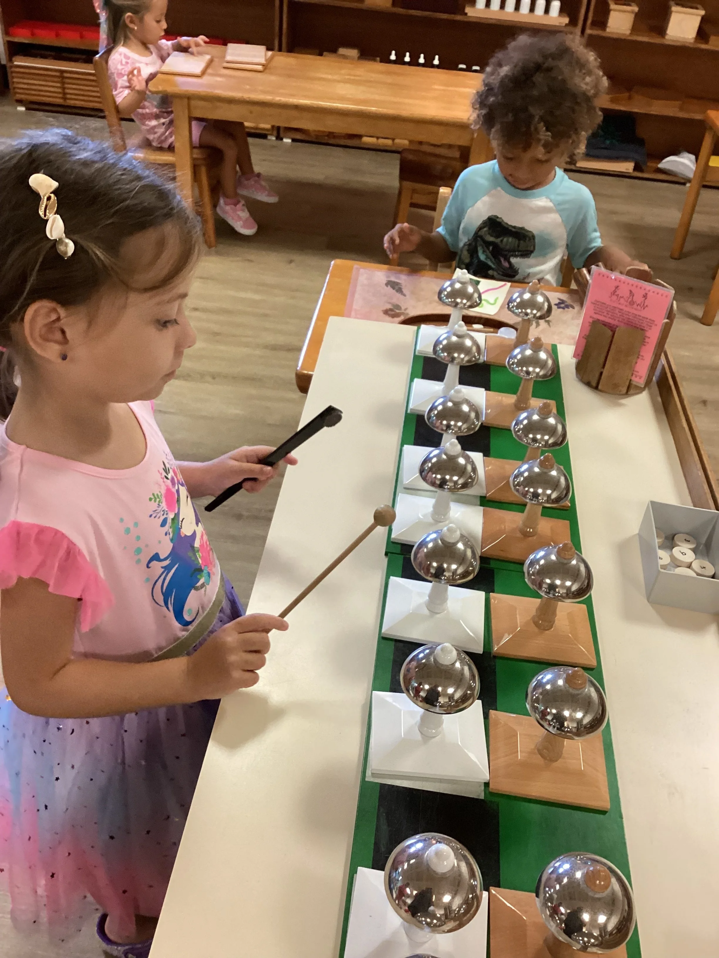 Two young girls playing a game of chess with metallic and wooden pieces on a small green chessboard in a classroom or play area.