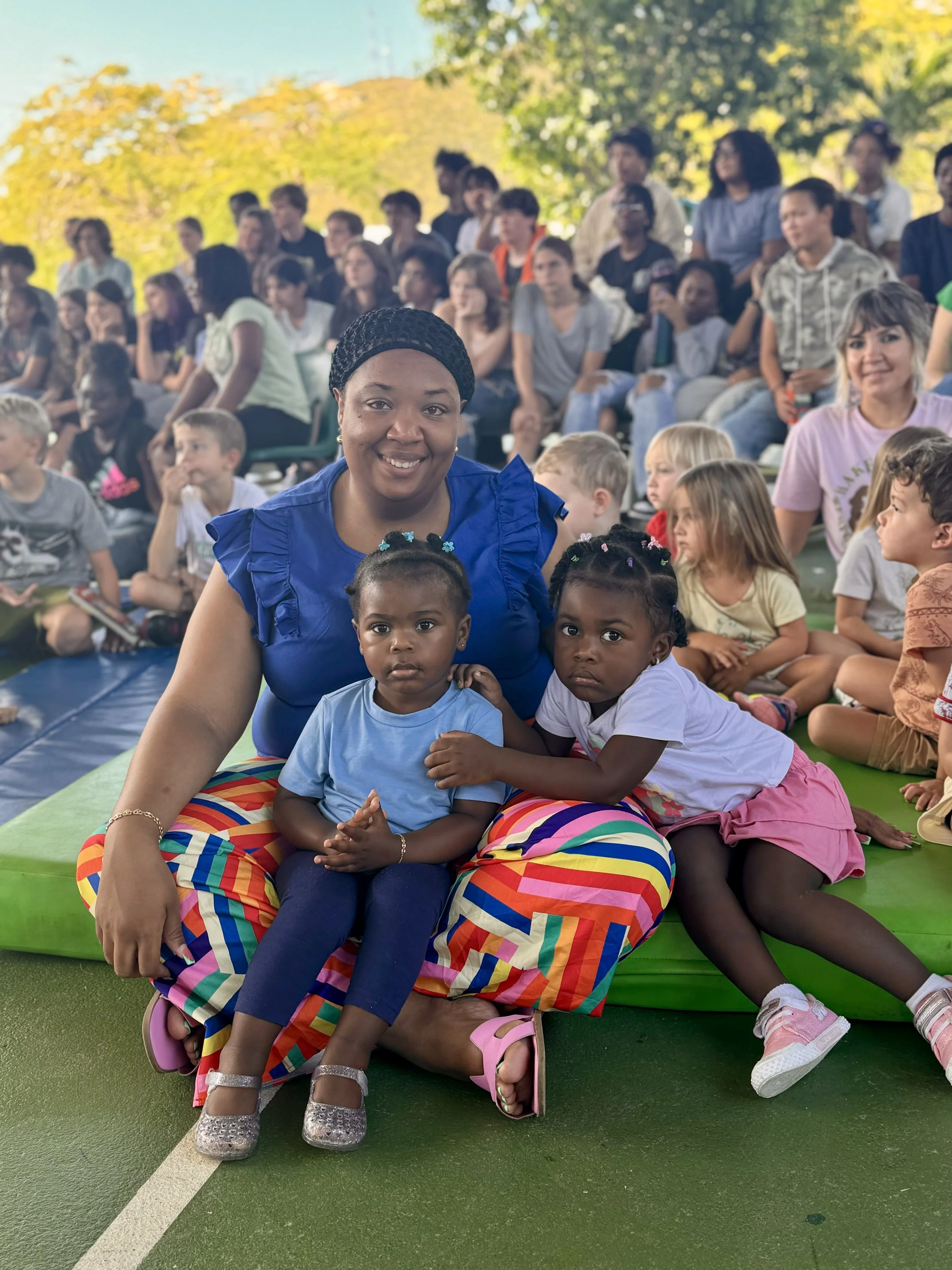 A woman sitting on the ground with two young girls, one in a light blue shirt and the other in a white shirt with pink shorts, surrounded by a crowd of children and adults outdoors on a sunny day.