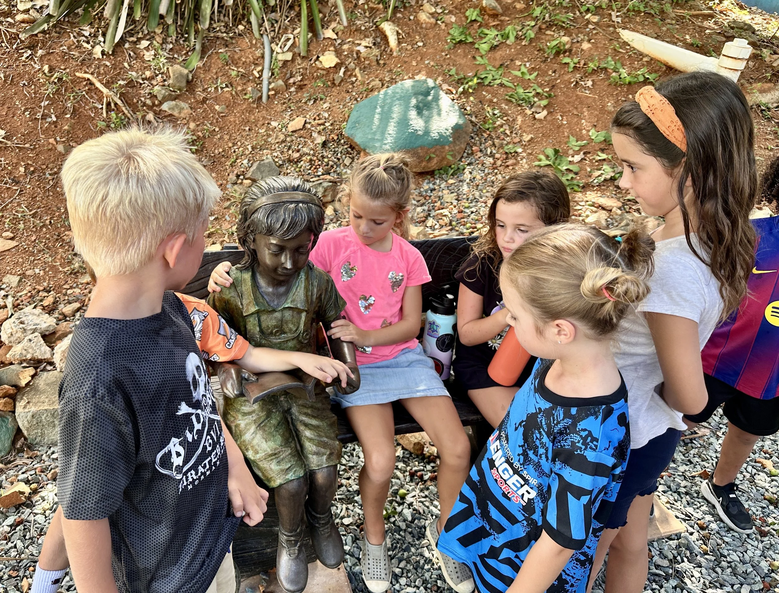 Group of young children gathered around a seated bronze statue of a girl reading a book outdoors.