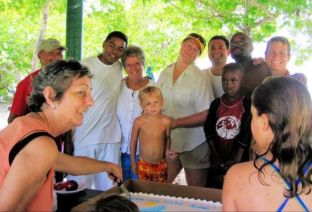 A group of people, including children and adults, gathered outdoors around a table with a cake, smiling and celebrating.