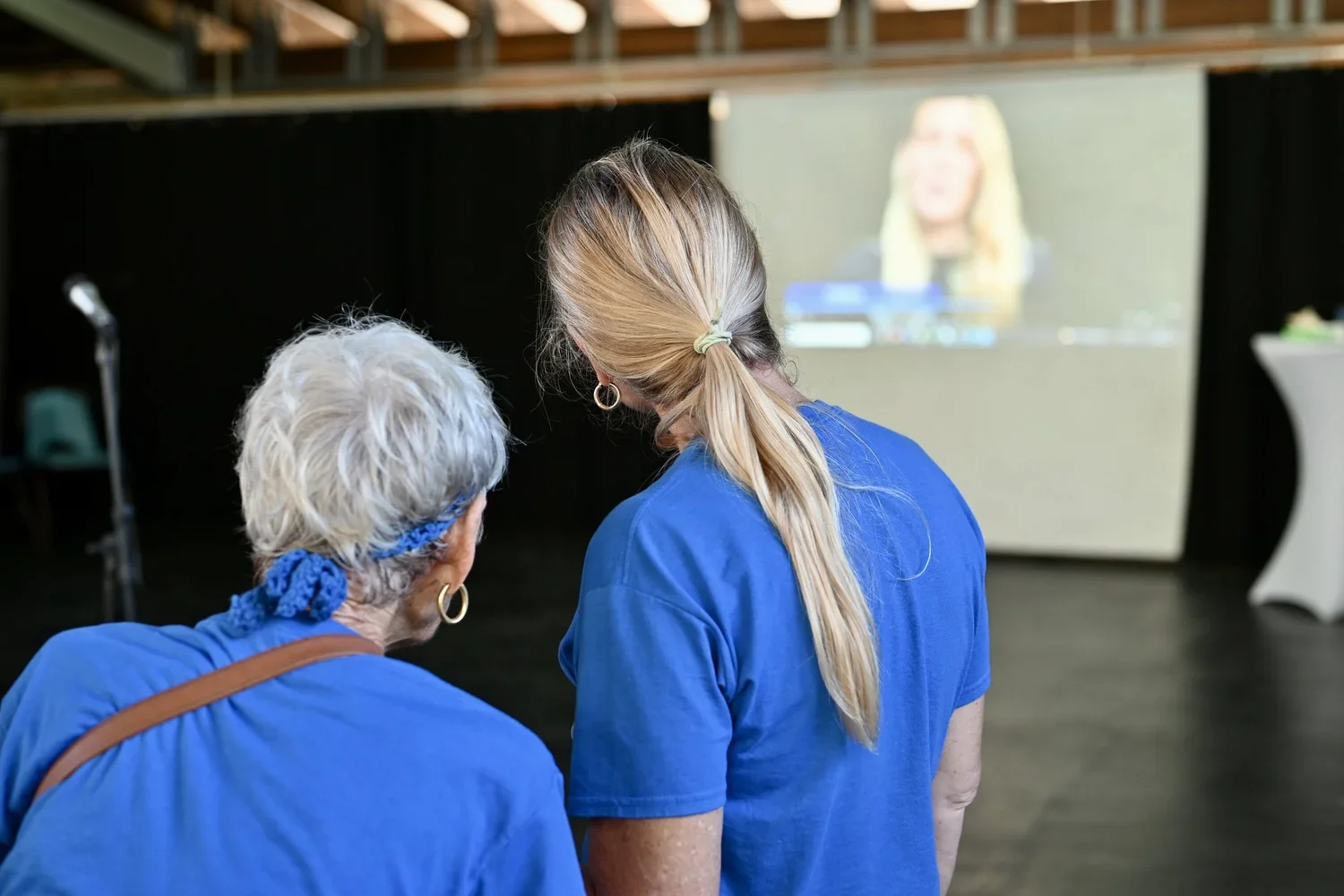 Two women, one older with white hair and the other younger with blonde hair, wearing blue shirts, watching a woman on a video call or presentation projected on a screen.