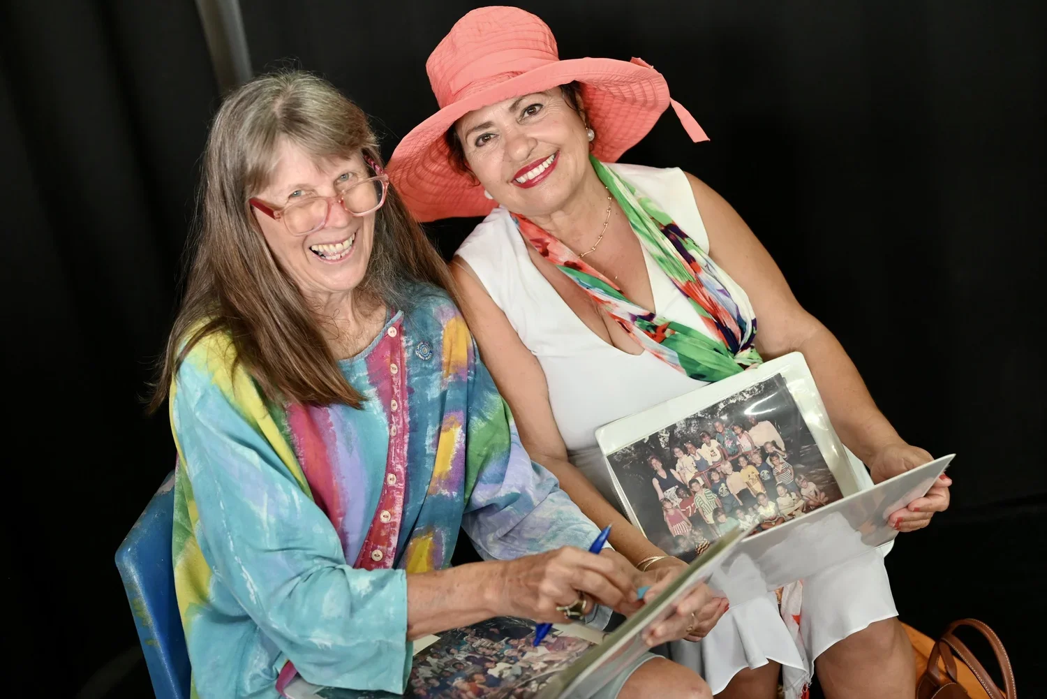 Two smiling women sitting together, one wearing a colorful tie-dye shirt and the other in a white dress with a large pink sunhat, holding a photo album showing a group picture.