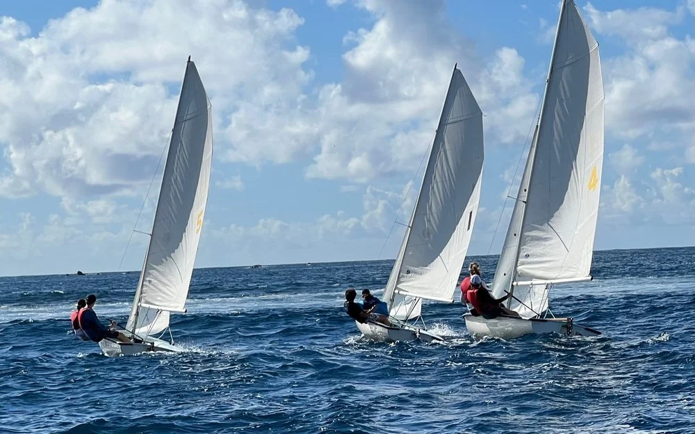 Three small sailboats with white sails racing on the open blue ocean under a partly cloudy sky.