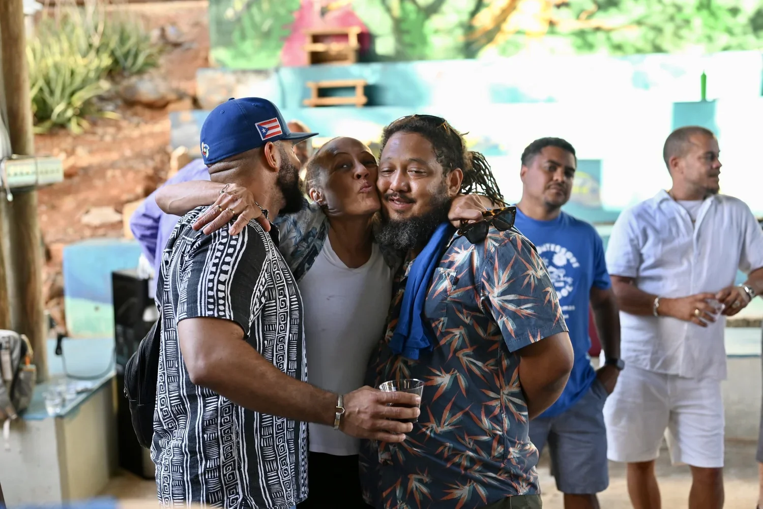 Group of friends hugging and socializing at a casual outdoor gathering, with some holding drinks and relaxing, in a colorful and lively setting.