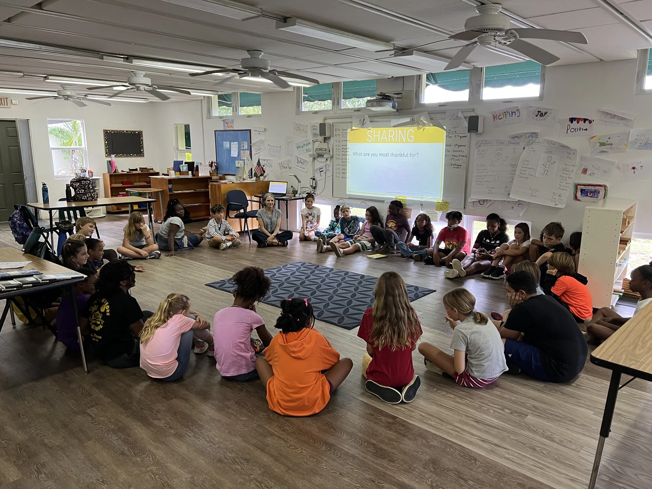 A classroom with children sitting on the floor in a large circle, participating in a group activity. A teacher is sitting among them. A projector screen displays the word "SHARING" and the question "What are you most thankful for?" The classroom has bright windows, posters, and drawings on the walls.