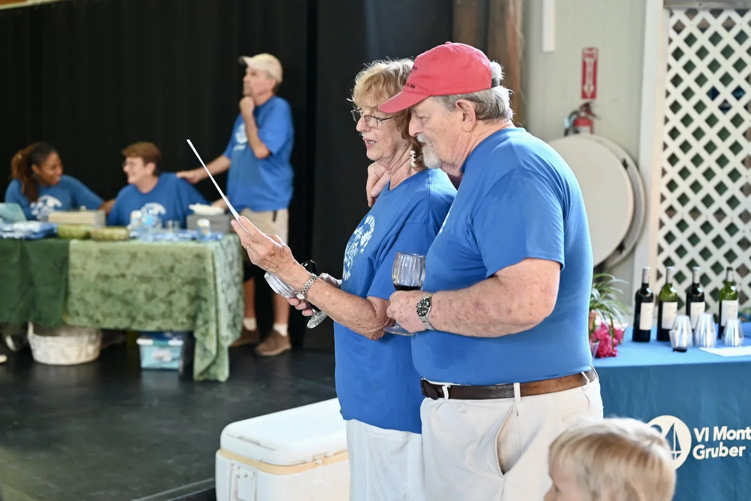 An elderly couple dressed in blue shirts is standing close together, reading a document at a social gathering. The woman is holding a wine glass, and the man is wearing a red cap and also holding a wine glass. In the background, there are people sitt