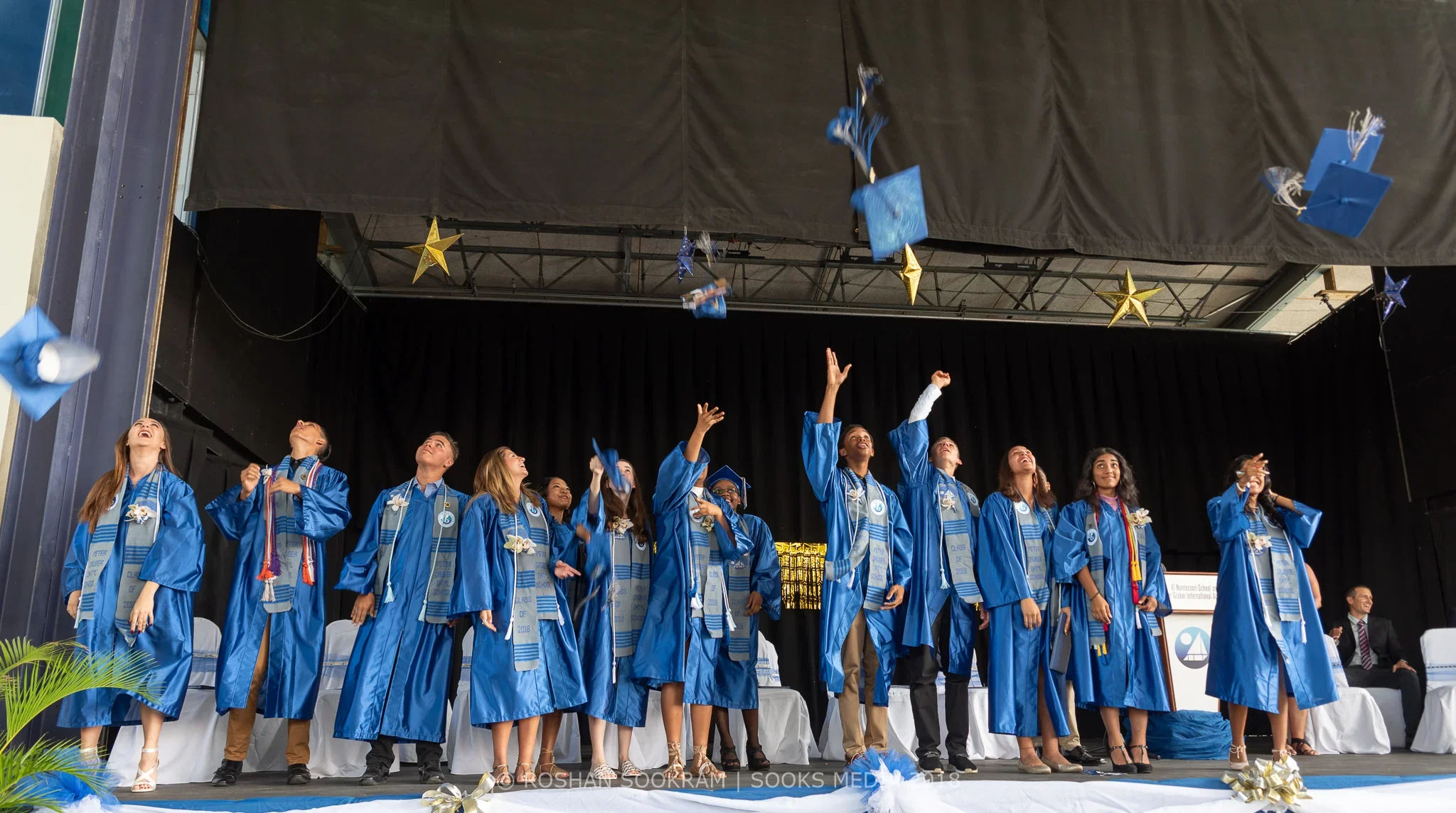 A group of graduates in blue caps and gowns celebrating on stage during a graduation ceremony, throwing their caps in the air.