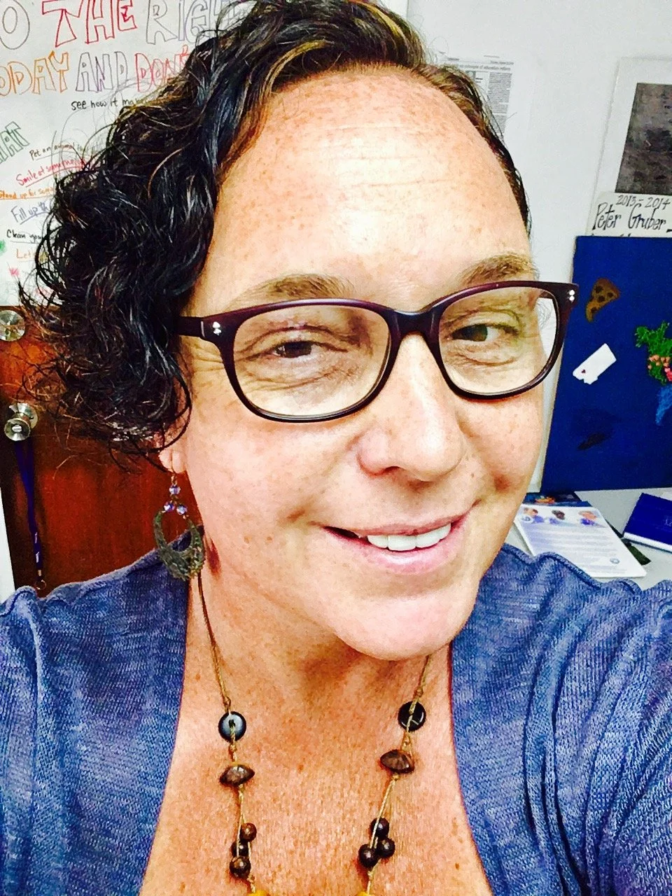 Close-up of a woman with short, curly black hair, wearing glasses, a blue top, large earrings, and a beaded necklace, smiling indoors with a colorful poster and whiteboard in the background.