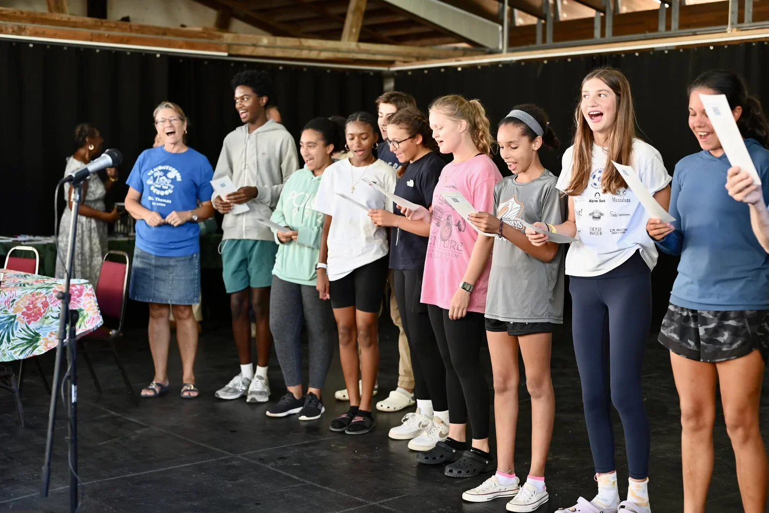 Group of young girls and women standing on a stage, reading from papers, with a black curtain background, during a presentation or performance.