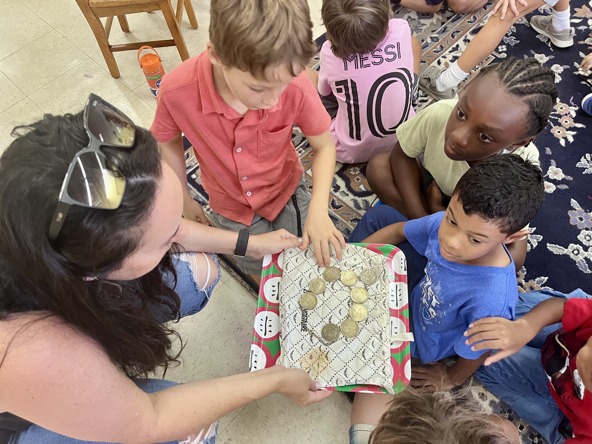 A woman shows children coins on a tray during an activity. The children are sitting on the floor, observing the coins closely.