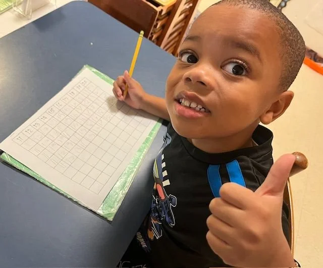 A young boy sitting at a table with a blank chart or calendar, holding a yellow pencil in one hand and giving a thumbs-up with the other, looking at the camera with a slight smile.