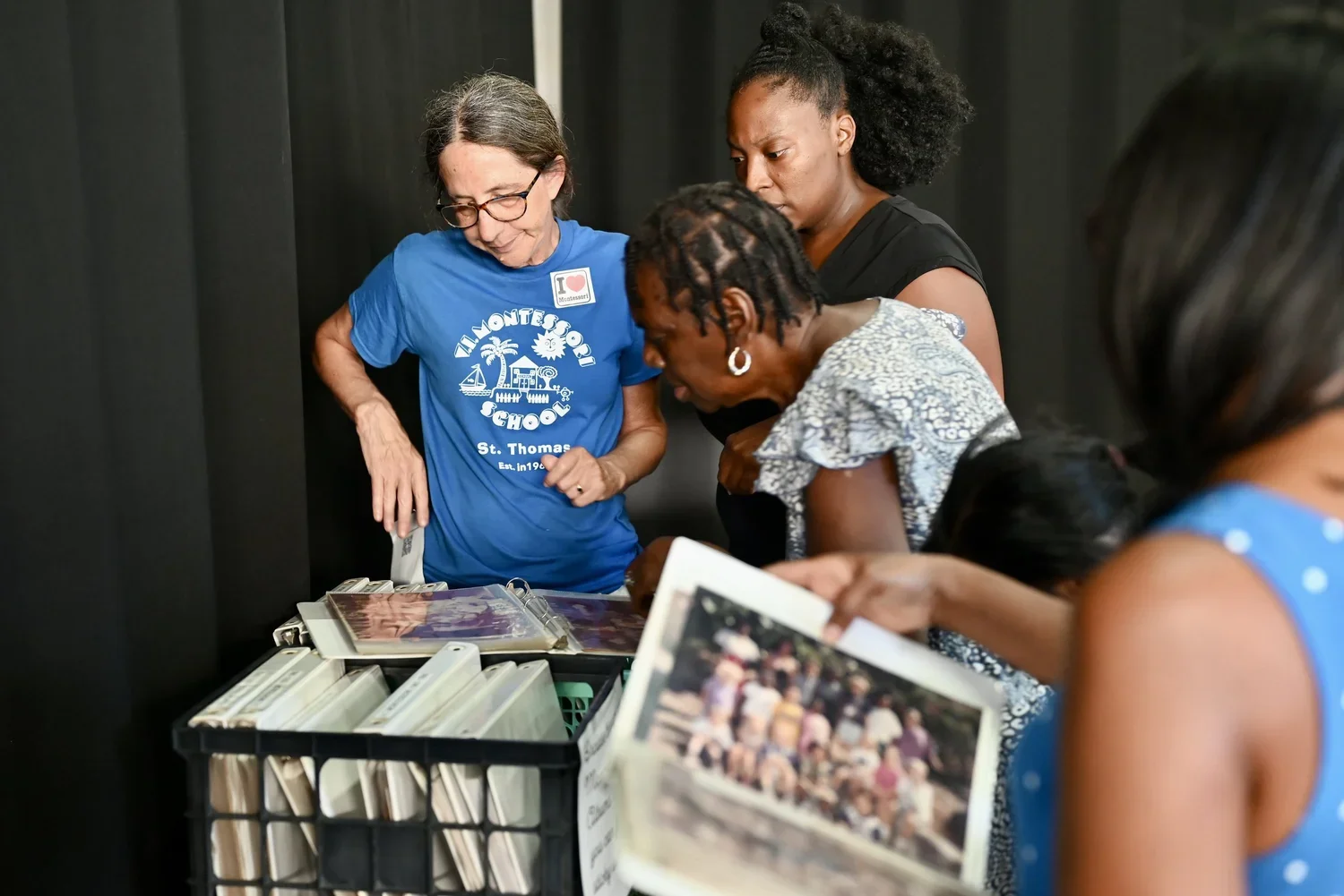 Four women gathered around a table looking at photographs or papers, with photo albums and pictures on the table, indoors with dark background.