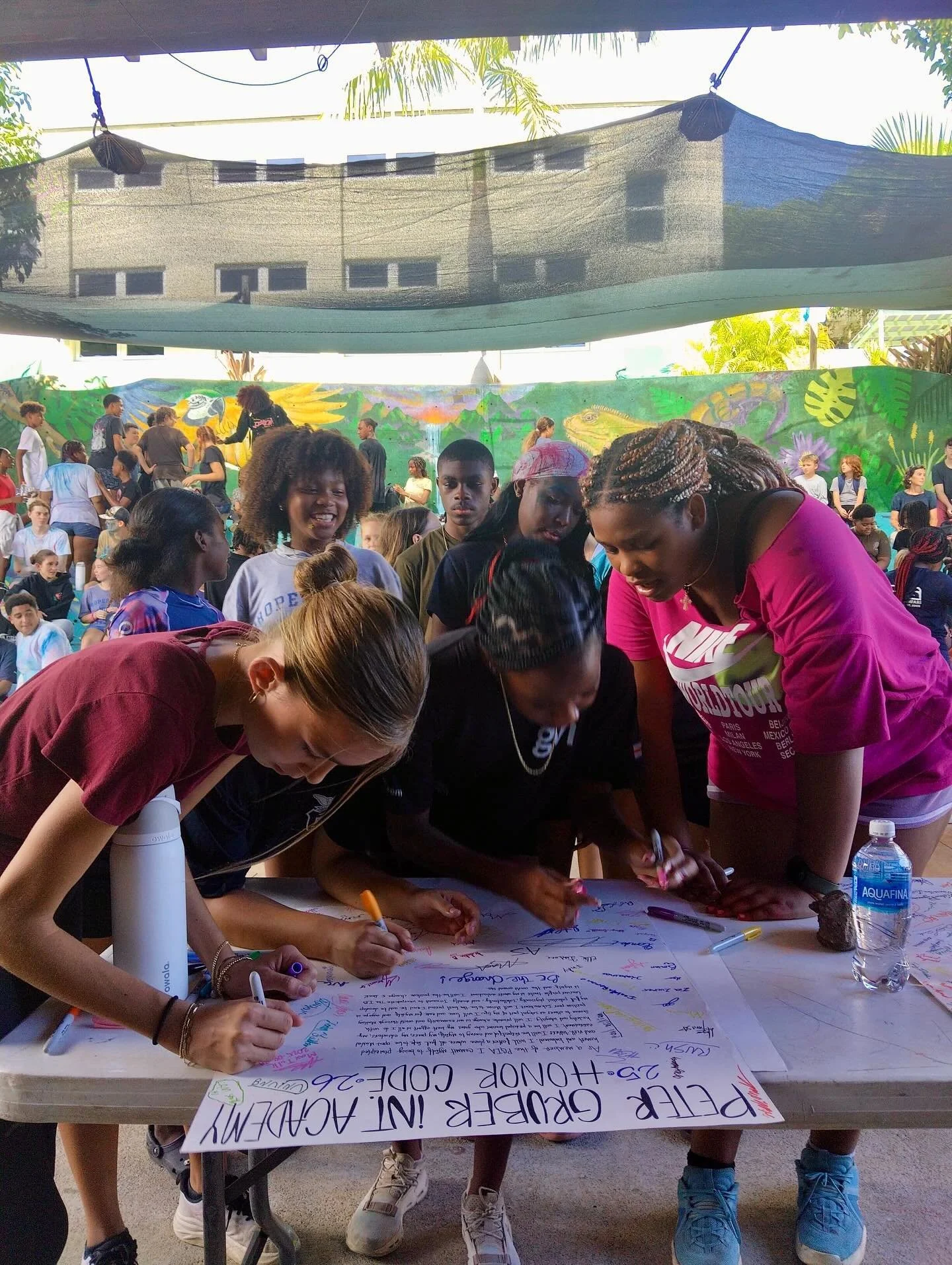 A group of young women signing a large poster at an outdoor event with many people in the background and a colorful jungle-themed mural on the wall.