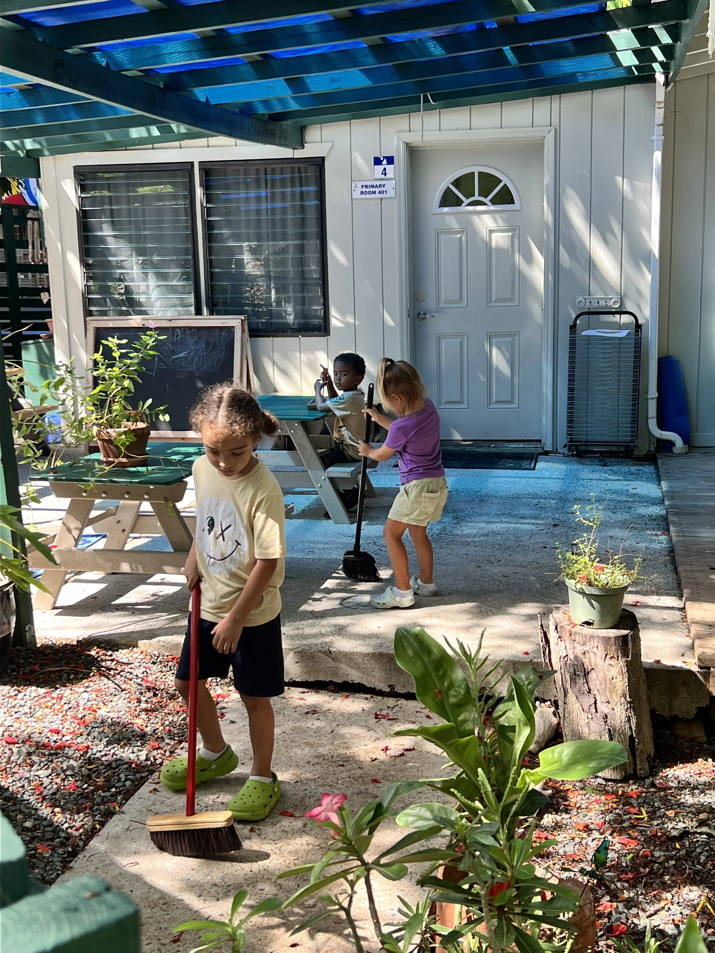 Three children playing outdoors in front of a building with a blue porch roof, one sweeping the ground, one sitting on a bench, and one standing with a broom, surrounded by potted plants and garden furniture.
