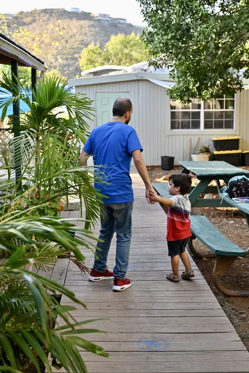 A man and a young boy holding hands and walking along a wooden path outside a small building, surrounded by greenery and outdoor furniture.