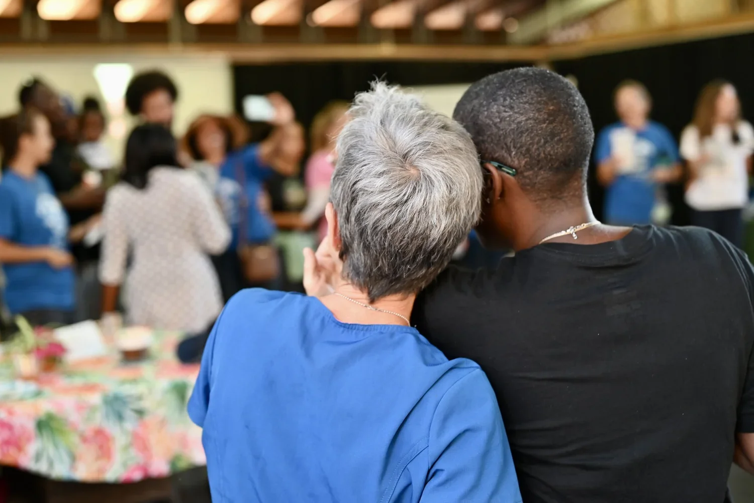 Two women, one with gray hair and wearing a blue shirt, and the other with short dark hair and wearing a black shirt, leaning close together while watching a group of people speaking at an event.