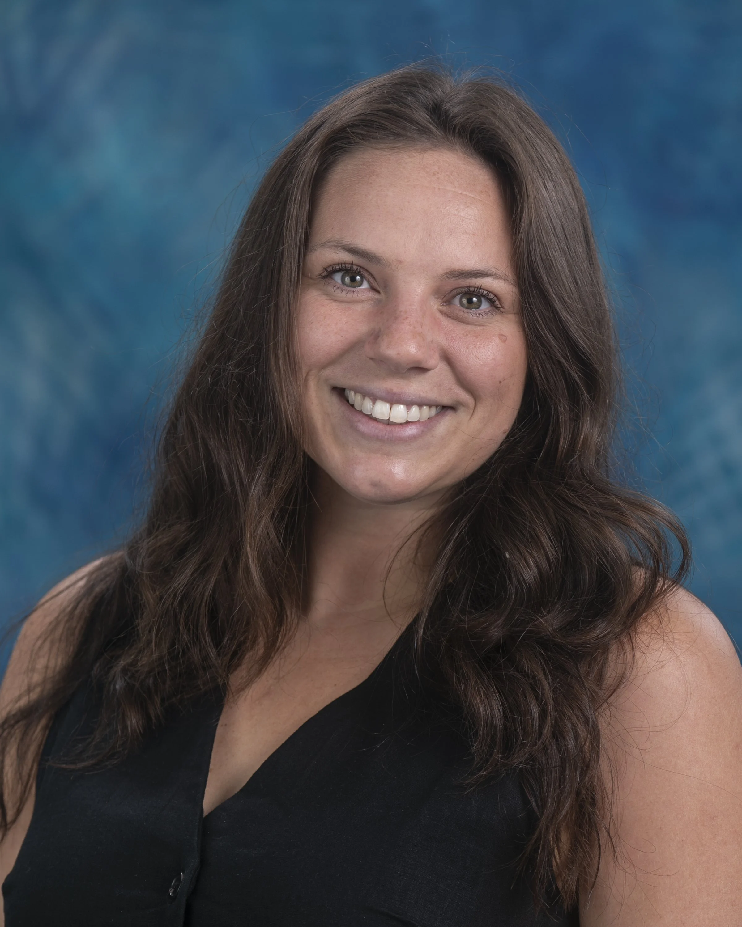 A woman with long brown hair smiling while crouching in a field of colorful tulips, including red, pink, yellow, and white flowers.