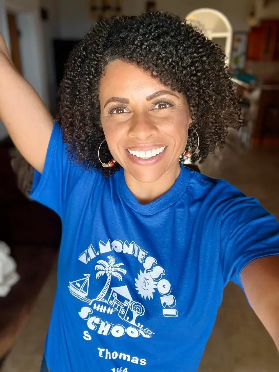 A smiling woman with dark curly hair taking a selfie, wearing a blue T-shirt that says 'Ti. Monte School' with a graphic of a house, boat, and trees, in a room with blurred background.