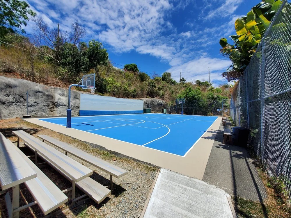 Outdoor basketball court with blue surface, surrounded by a chain-link fence, benches, and trees under a partly cloudy sky.