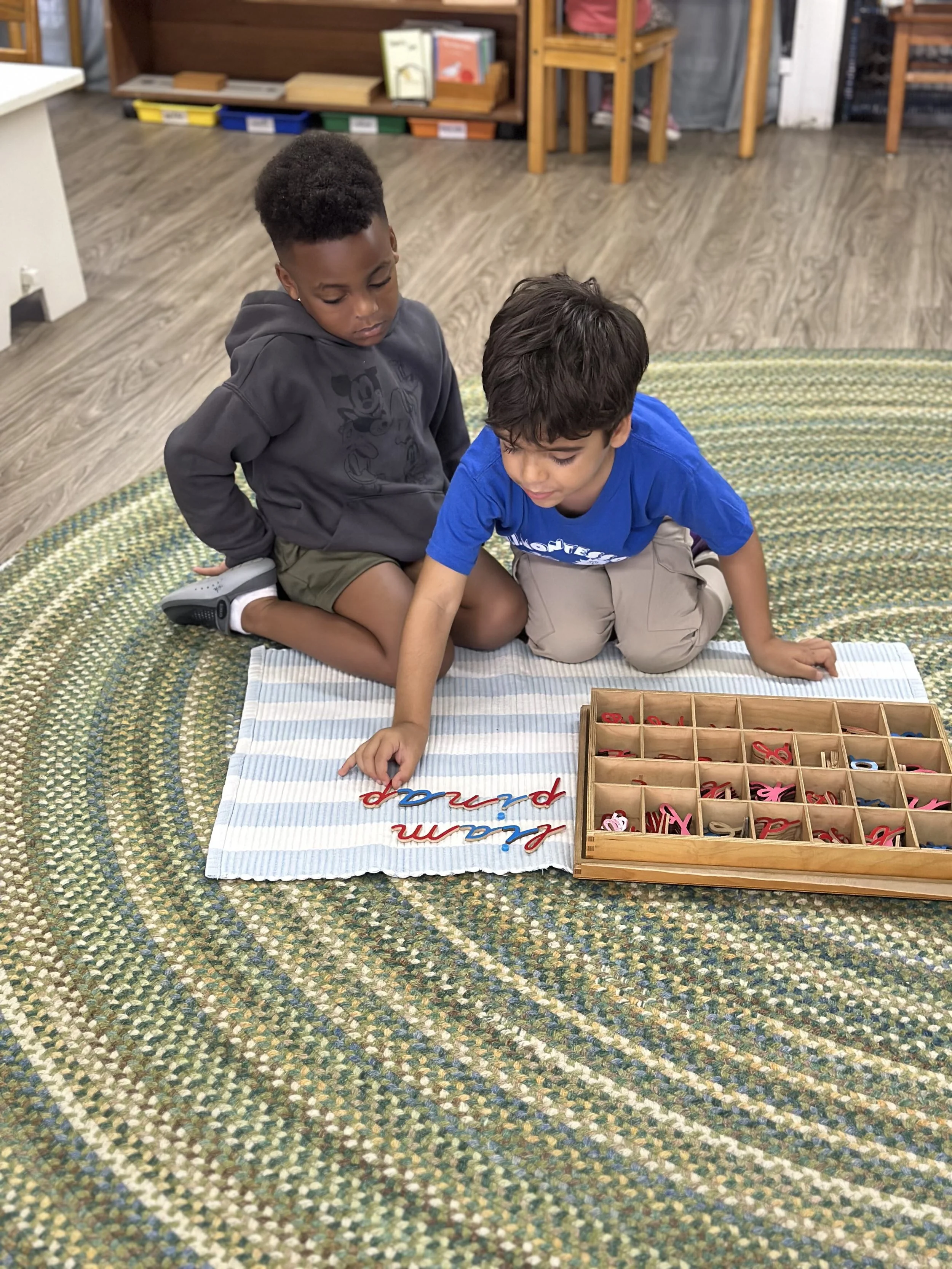 Two young boys sitting on a circular green patterned rug, engaging in a craft project with colorful letters and a wooden box of small items, in a room with wooden furniture and bookshelves.
