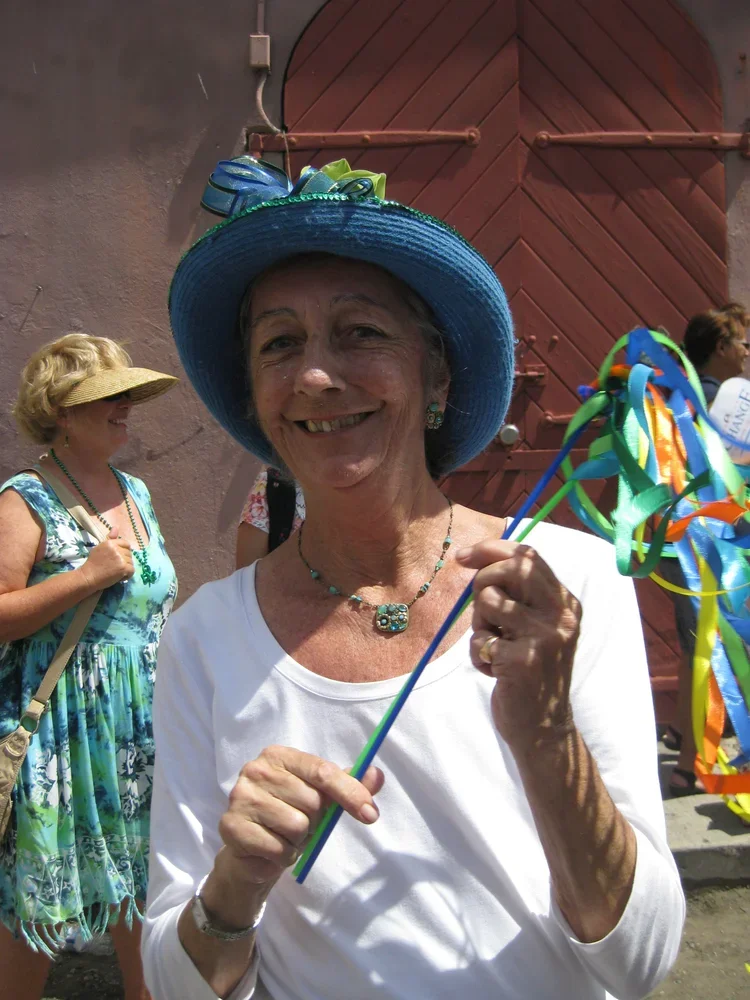 Smiling elderly woman wearing a blue hat with a green ribbon, holding colorful ribbon wands, at an outdoor celebration with other women in the background.