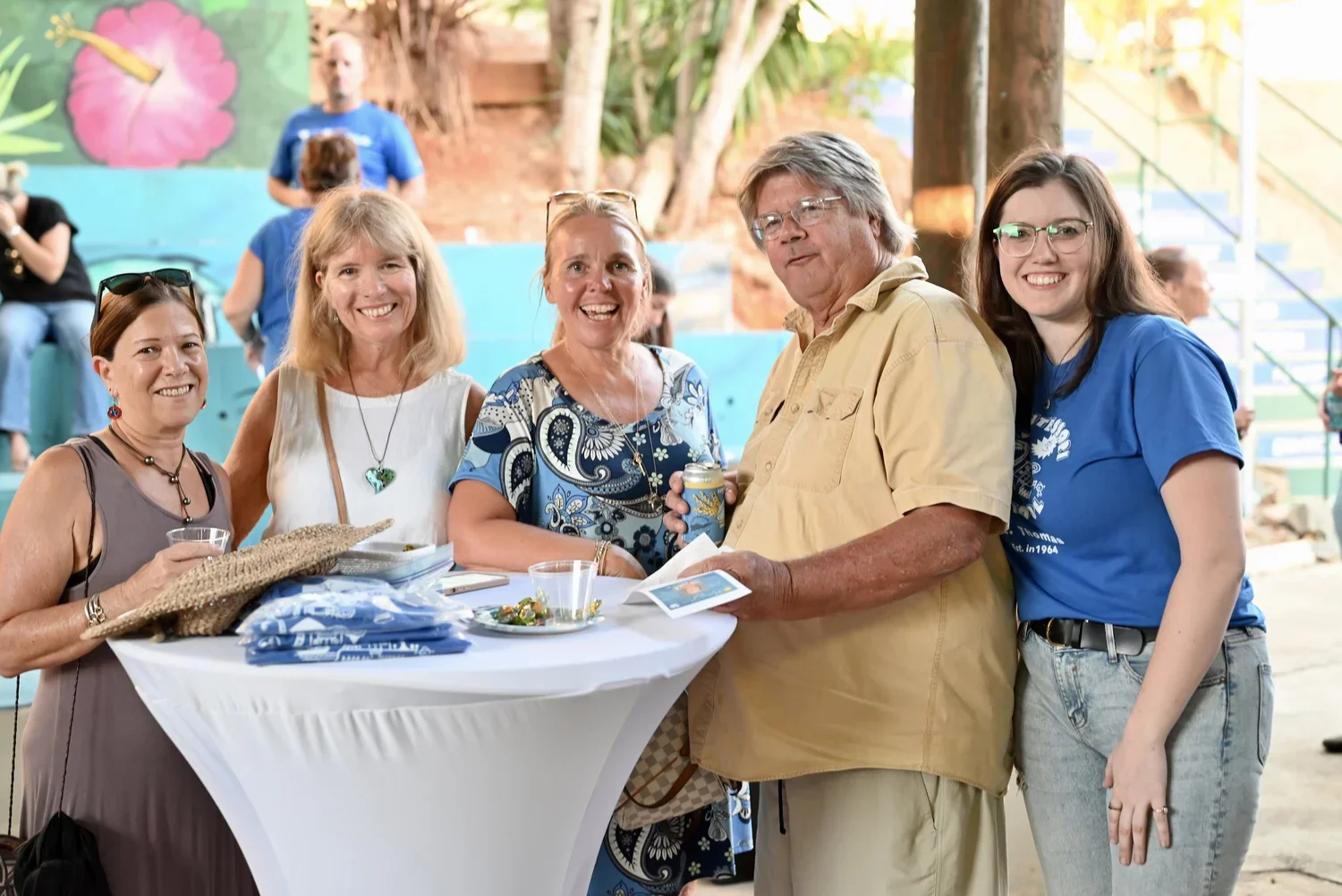 Group of five smiling women and one man gathered around a table at a social event, with some holding drinks. Background features colorful wall art and other people.
