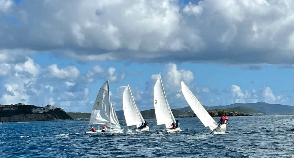 Four sailboats racing on the water with large white sails, under a partly cloudy sky, with land and hills in the background.