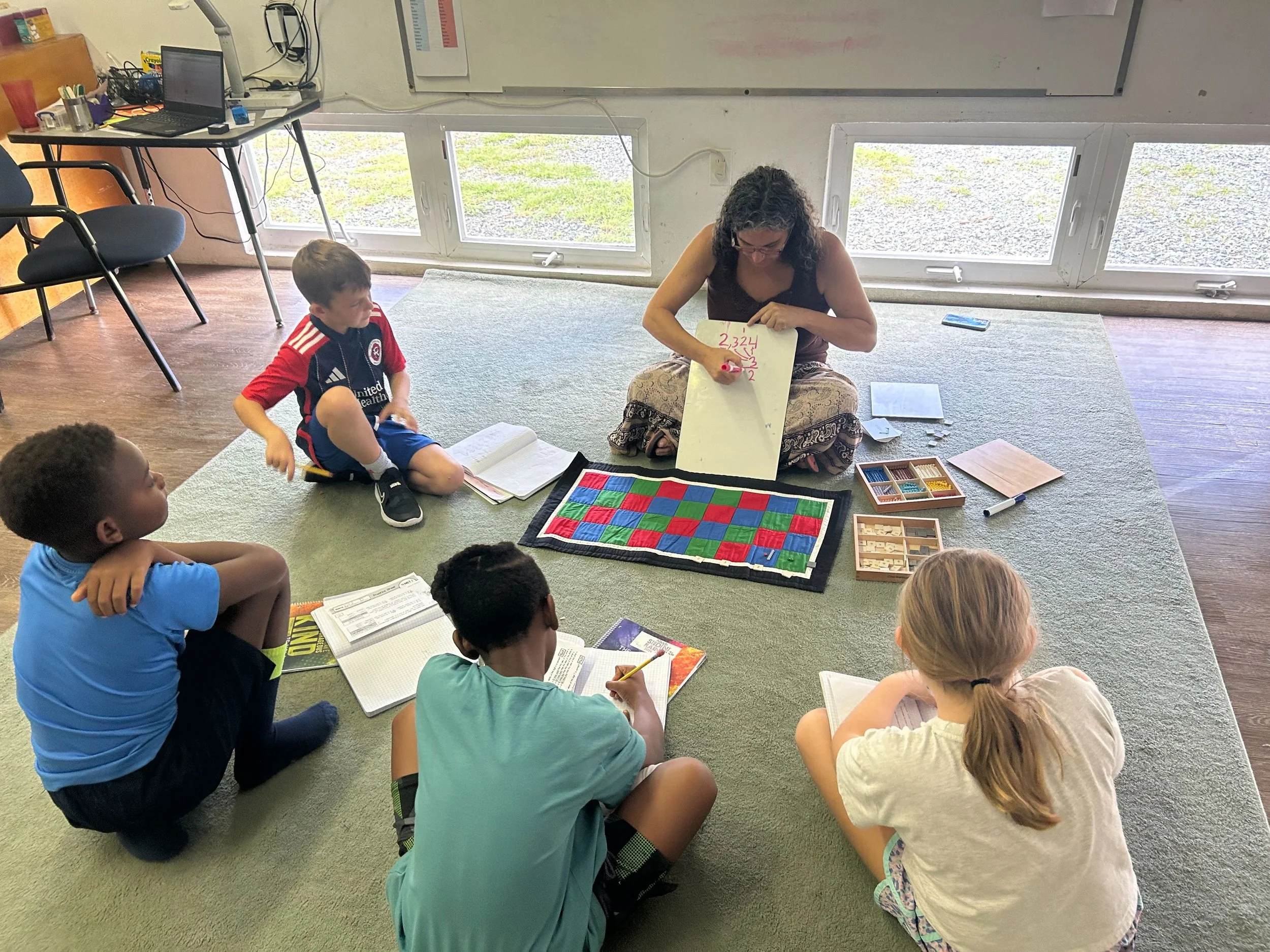 A teacher sitting on the floor teaching a math lesson to five children who are seated around her on carpet. The teacher uses a whiteboard with written math problems, and the children have open books and notebooks in front of them.