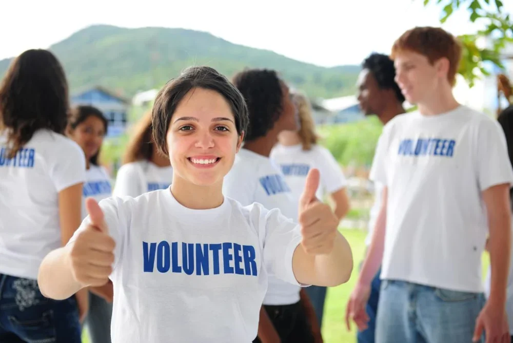 A young woman smiling and giving a thumbs-up while wearing a white volunteer shirt at an outdoor community event, with other volunteers in the background