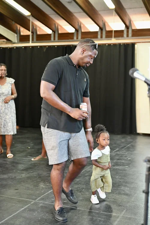 A smiling young man holding a microphone, walking on a stage with a small girl, another woman standing in the background, in a venue with wooden beams and black curtains.