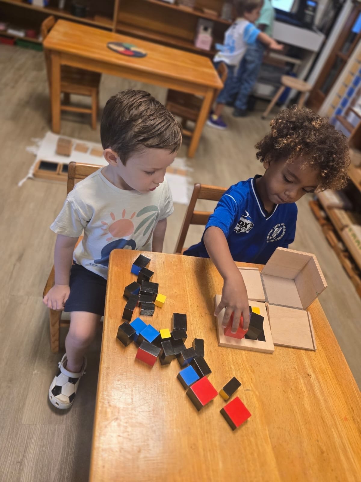 Two young boys at a classroom table playing with colorful wooden blocks and a wooden storage box, with one boy wearing a white t-shirt and the other in a blue sports jersey.