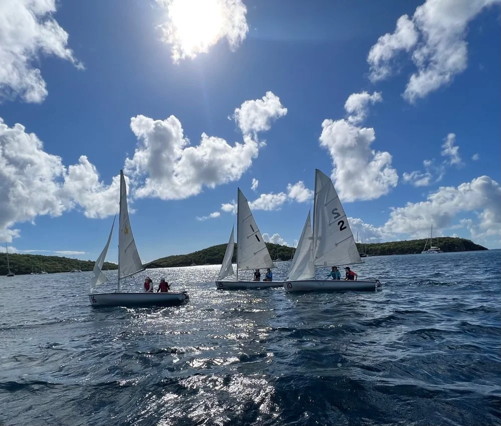 Three small sailboats with people sailing on the ocean, with a bright blue sky and white clouds overhead, and a distant landmass in the background.