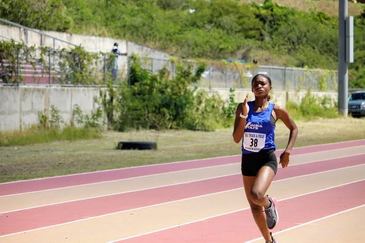 Female track athlete running on a red and beige outdoor track, wearing a blue sleeveless top and black shorts, with a race bib numbered 38.