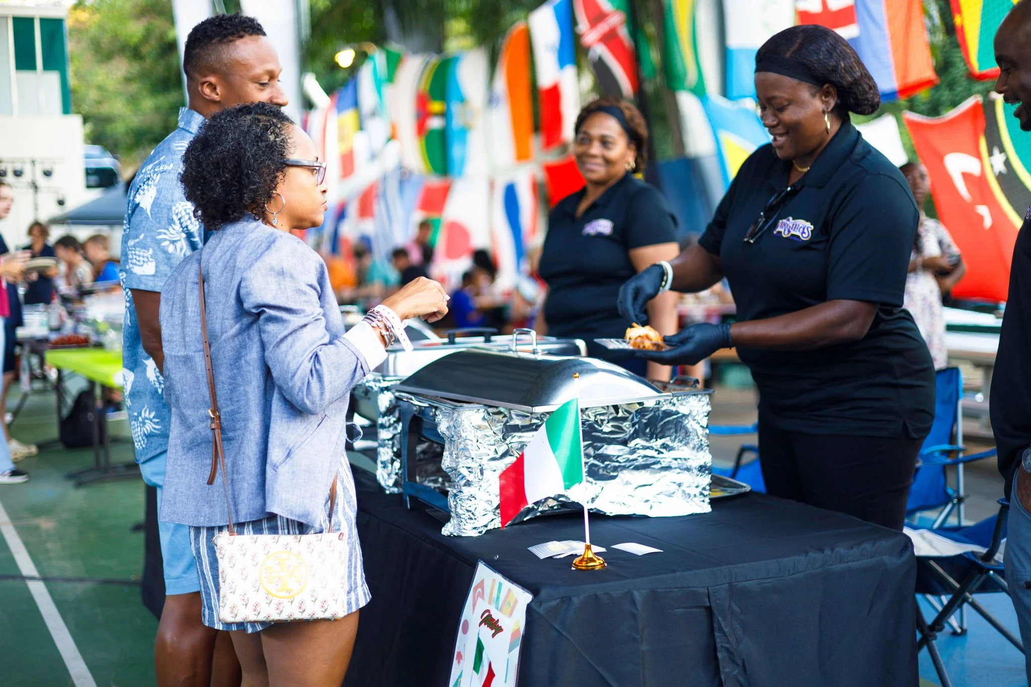 People at an outdoor food booth with an Italian flag, serving food at a festival or event with flags and banners in the background.