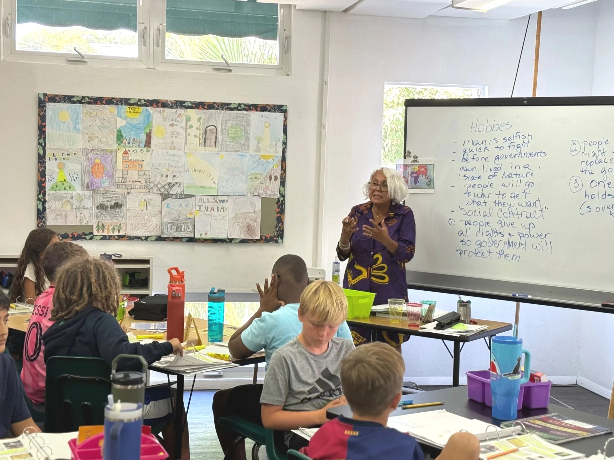 A classroom with students sitting at desks, attentively listening to a teacher standing at the whiteboard. The teacher is an older woman with gray hair, wearing a purple shirt and a patterned sash. The whiteboard behind her has notes about Hobbes, including points about his views on human nature and government. There is a large window at the back. The back wall displays colorful student drawings and writings about summer.