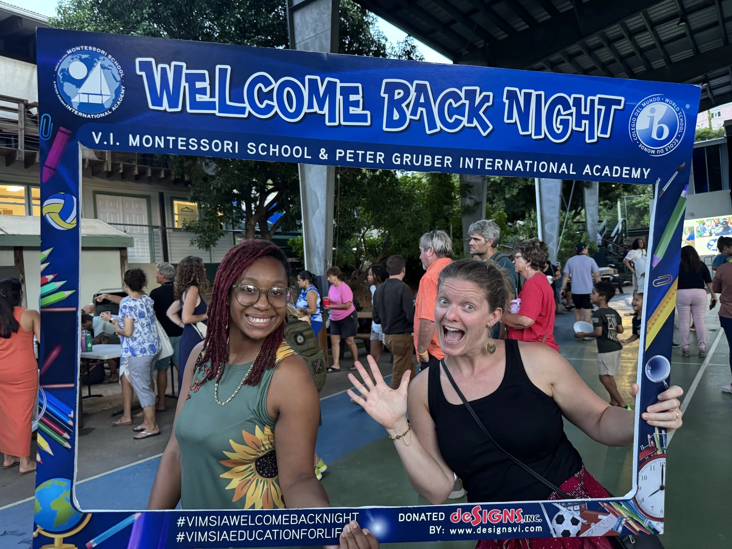 Two women smiling and waving at the camera with a group of people behind them at a welcome back night event at V.I. Montessori School and Peter Gruber International Academy.