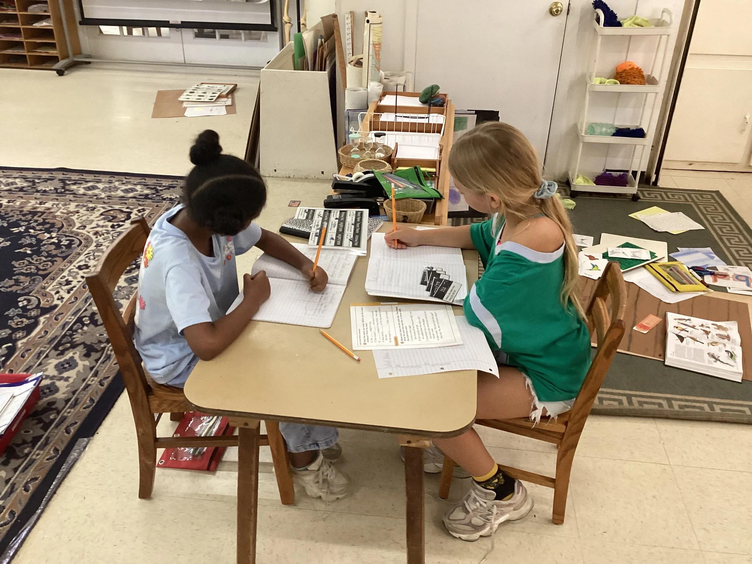 Two young girls sitting at a small table working on notebooks with school supplies in a classroom.