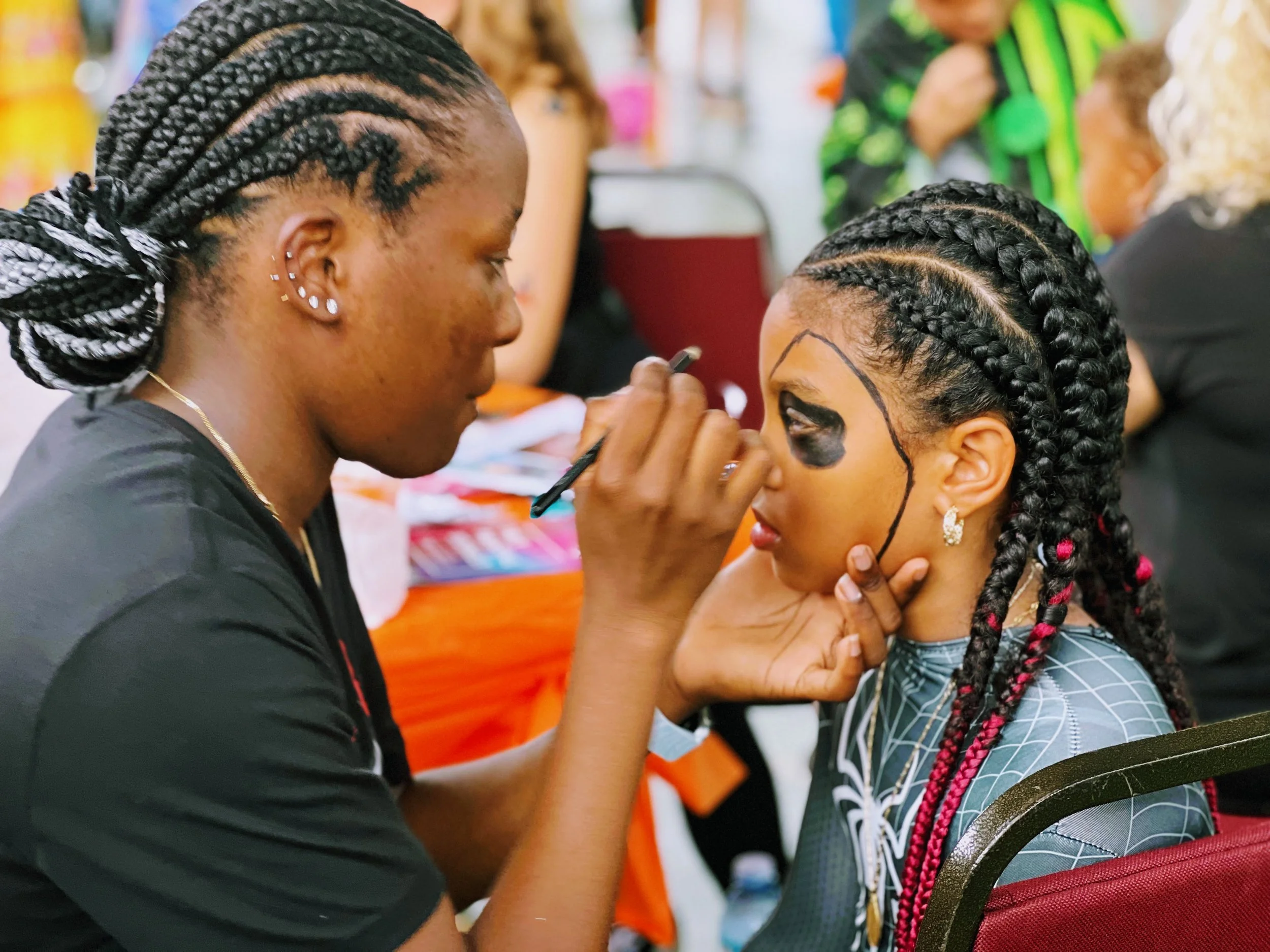 A woman with braided hair and earrings is painting a spooky mask with black makeup on a young girl's face, who has braided hair with red highlights, at a Halloween event.