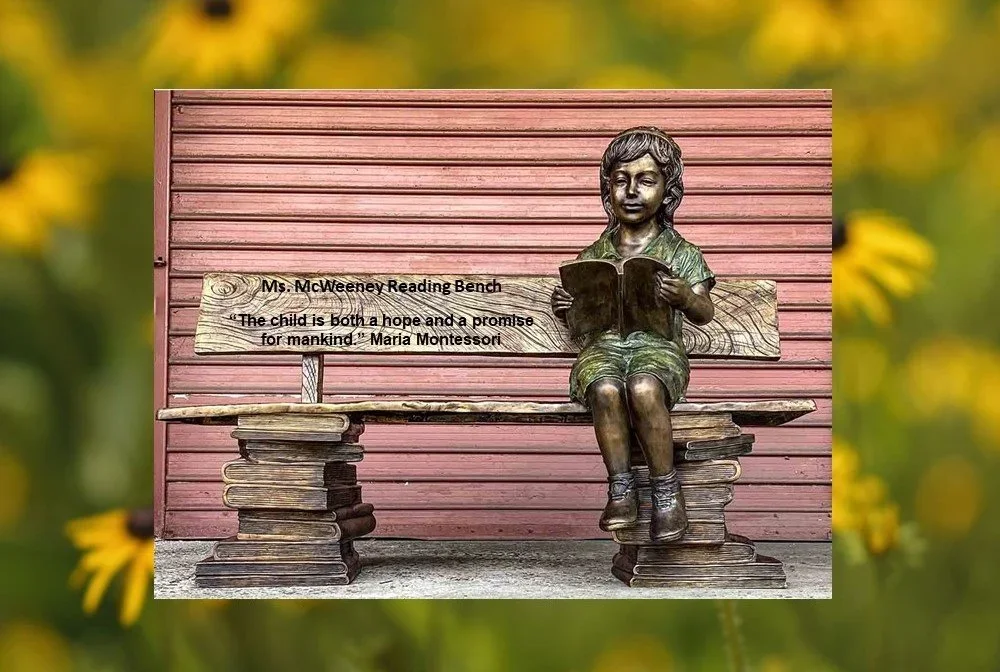 Bronze sculpture of a young girl sitting on a bench, reading a book, with books forming the legs of the bench. A wooden plaque on the bench reads, 'Ms. McWeeney Reading Bench. "The child is both a hope and a promise for mankind" - Maria Montessori.'