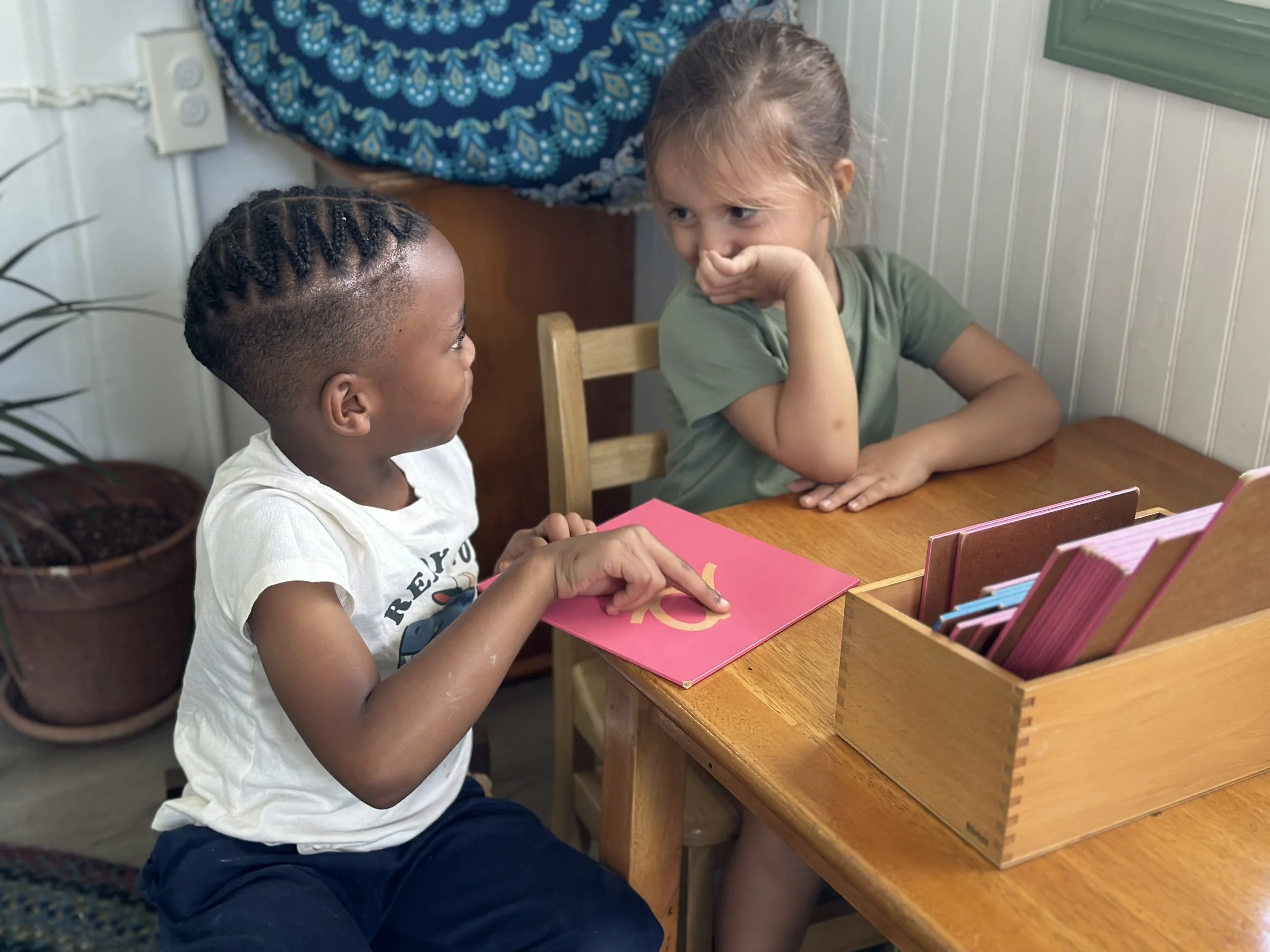 Two young girls sit at a wooden table, engaged in conversation. The girl on the left, with dark skin and braided hair, is pointing at a pink card. The girl on the right, with lighter skin and light brown hair, is resting her chin on her hand, listening thoughtfully.