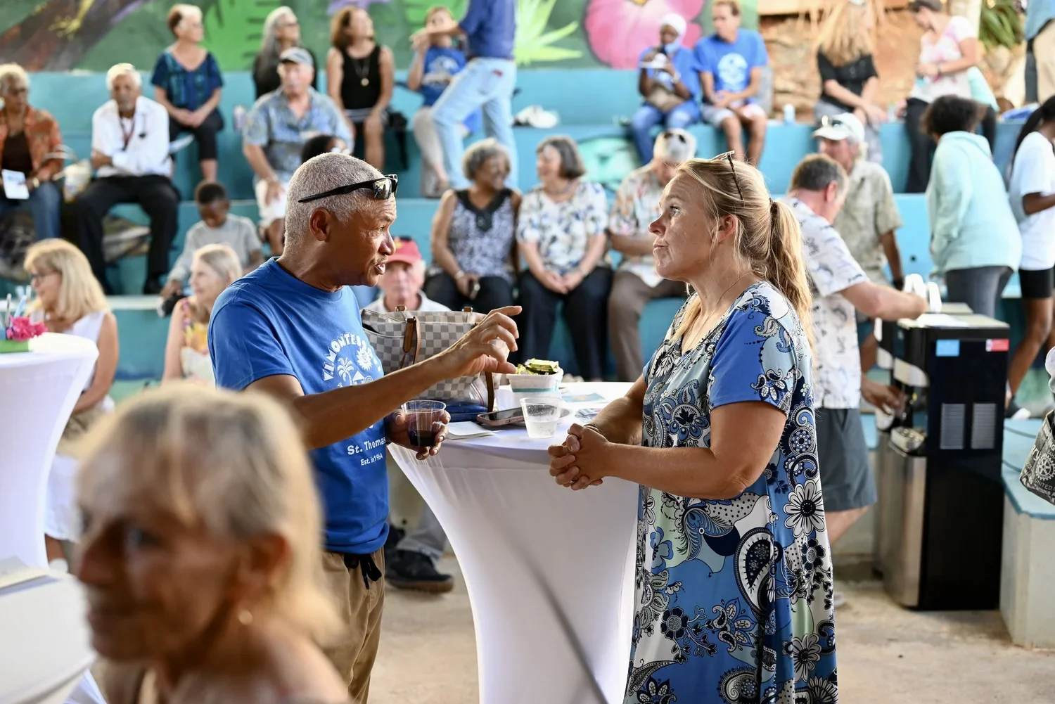 A man and a woman having a conversation at a social gathering, with multiple people sitting and standing in the background in an outdoor setting.