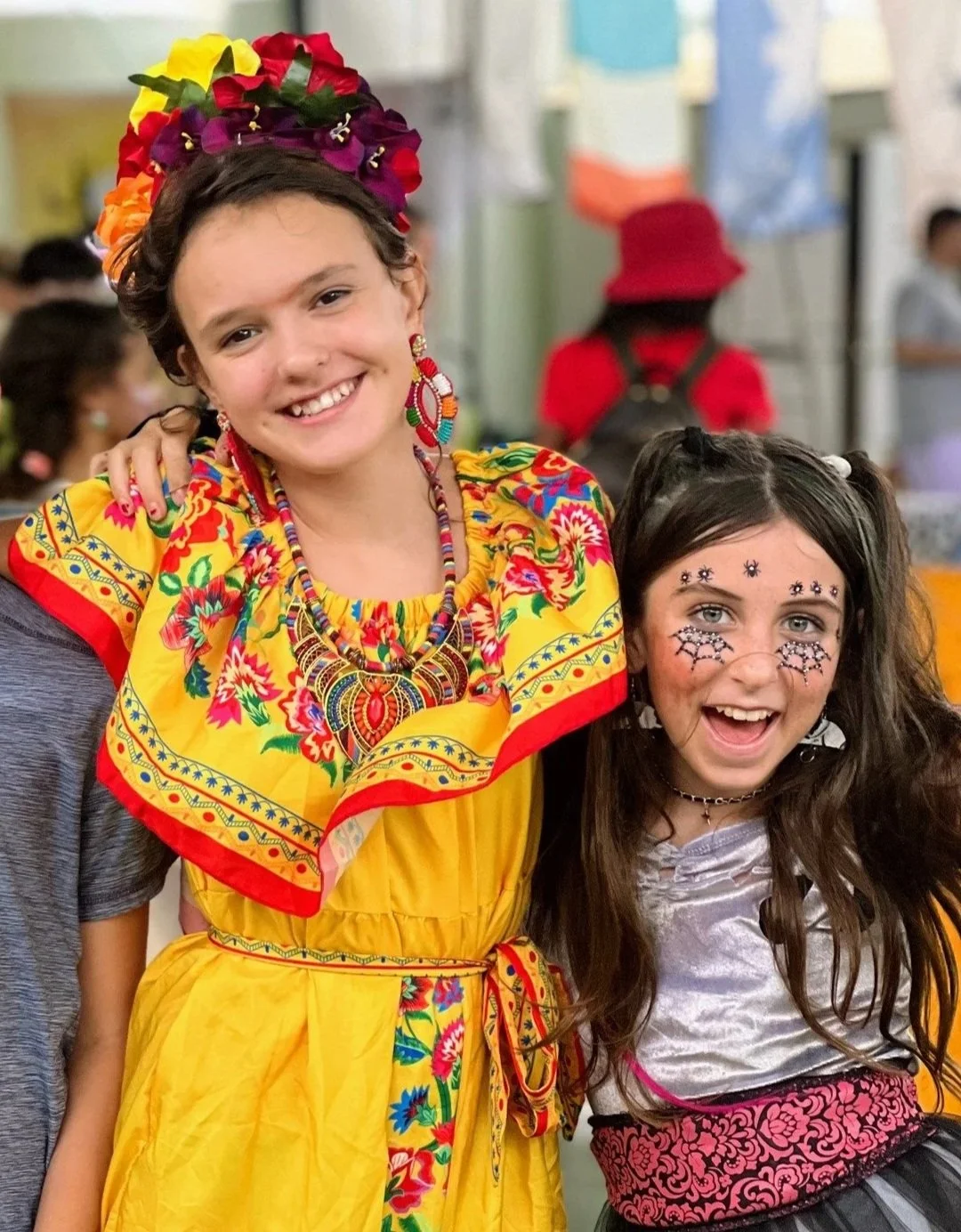 Two young girls in colorful costumes, smiling at a celebration. One girl wears a traditional floral dress and jewelry, with a headpiece made of flowers. The other girl has face paint decorated with Halloween designs and wears a fun dress. They are in a festive indoor setting.