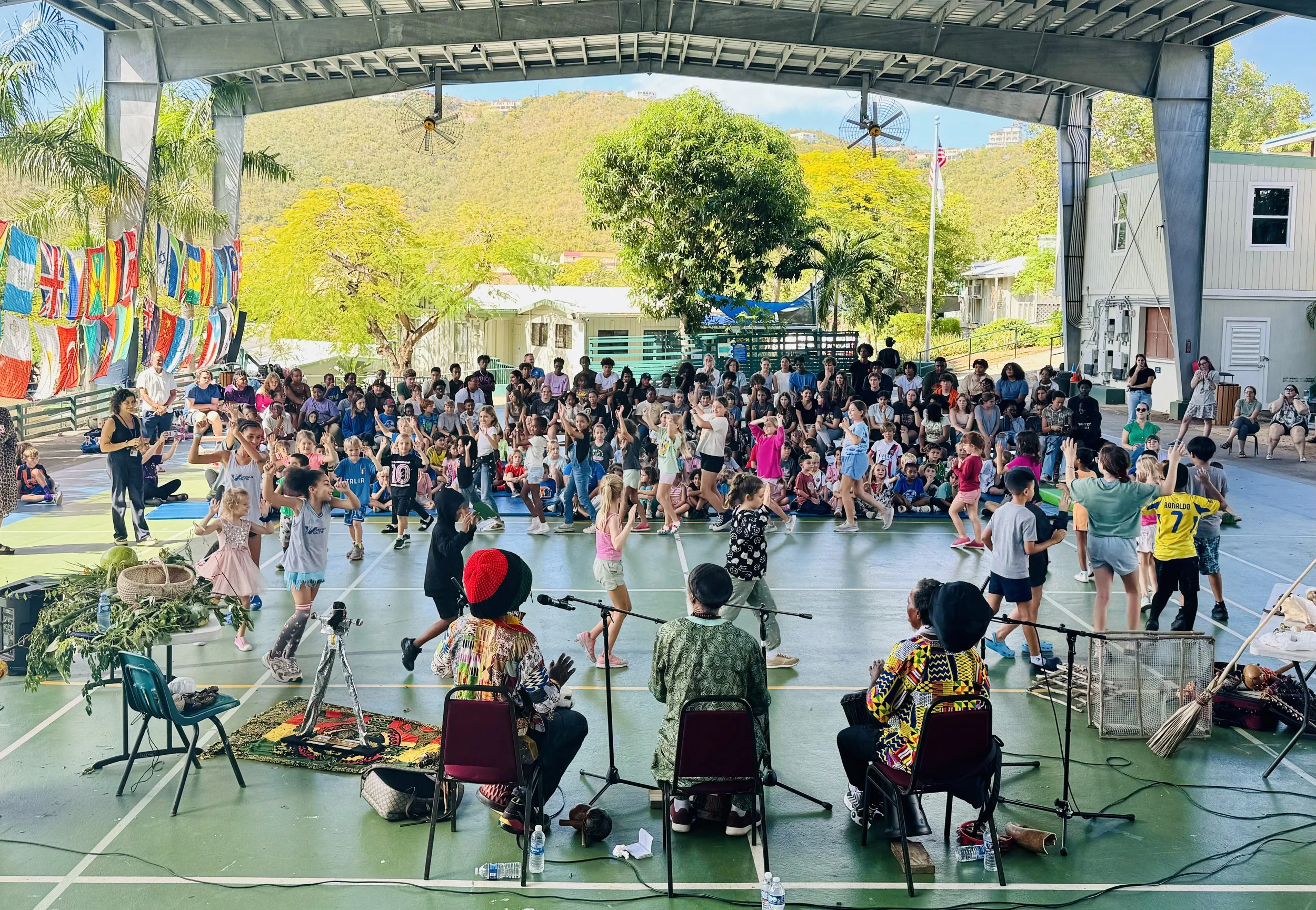 Large group of children and adults dancing and enjoying a performance in an open-air pavilion with a stage, surrounded by trees and mountains in the background.