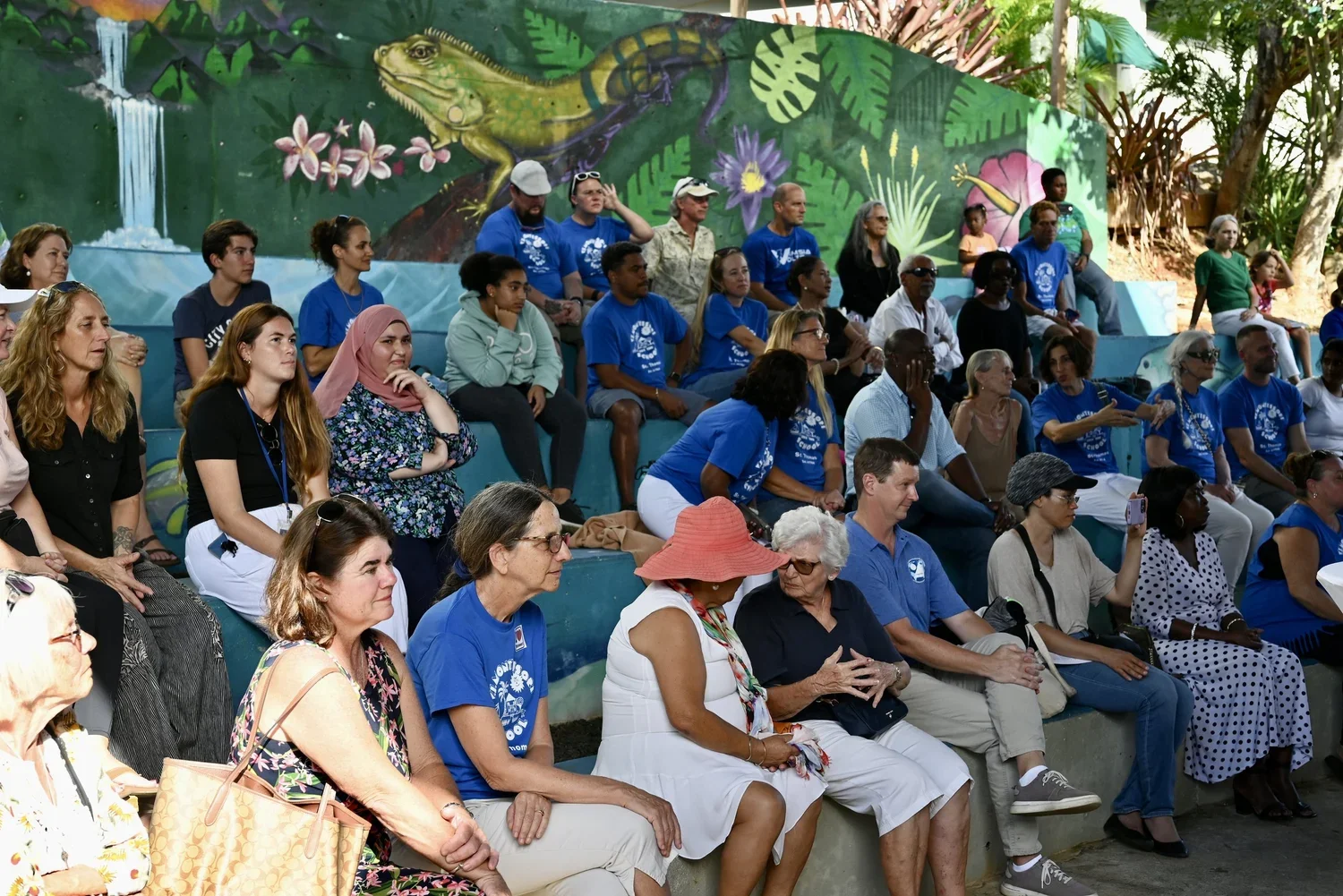 A diverse group of people sitting on tiered concrete seating, attending an outdoor event. The background features a colorful mural with a lizard, tropical plants, flowers, and a waterfall.